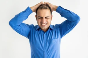 Hombre con camisa azul y expresión de preocupación o estrés, agarrándose la cabeza, lo que sugiere la angustia por la caída del cabello o la calvicie.
